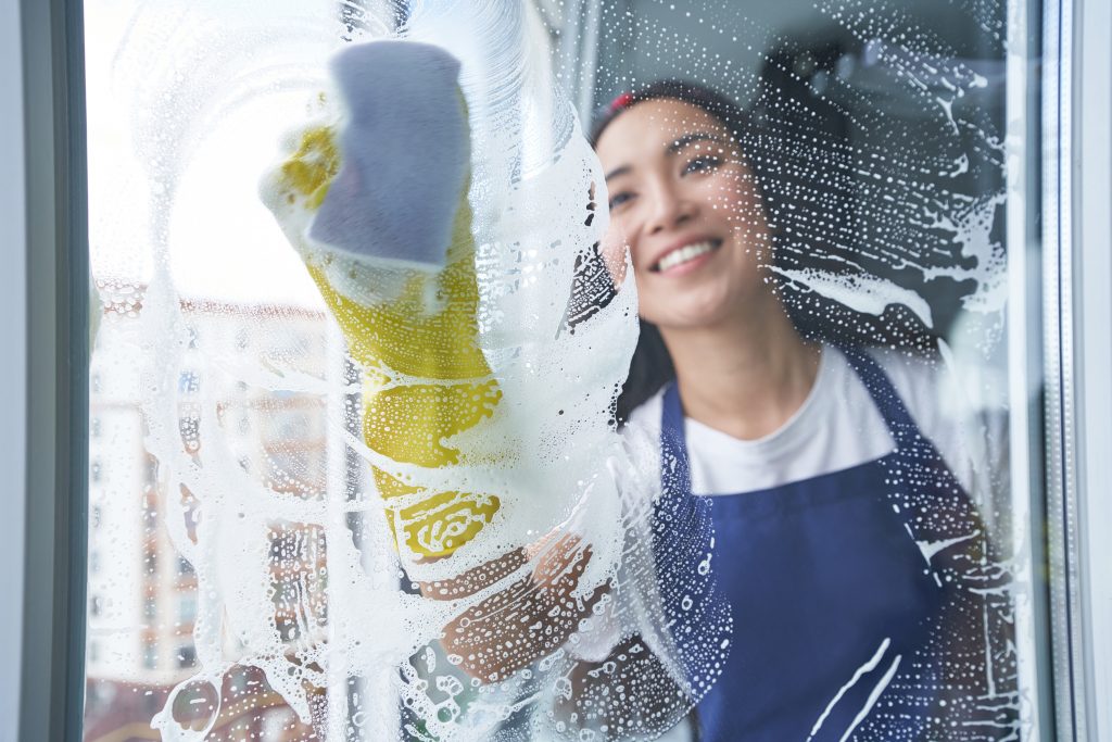 AdobeStock_392926234_©Friends Stock_Best cleaning service. Cheerful young woman smiling while cleaning the window, glass surface using sponge